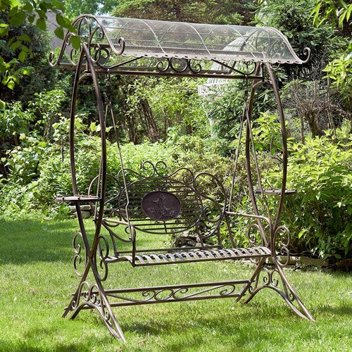 Ornate metal swing bench with scrollwork and a mesh canopy stands in a leafy garden.