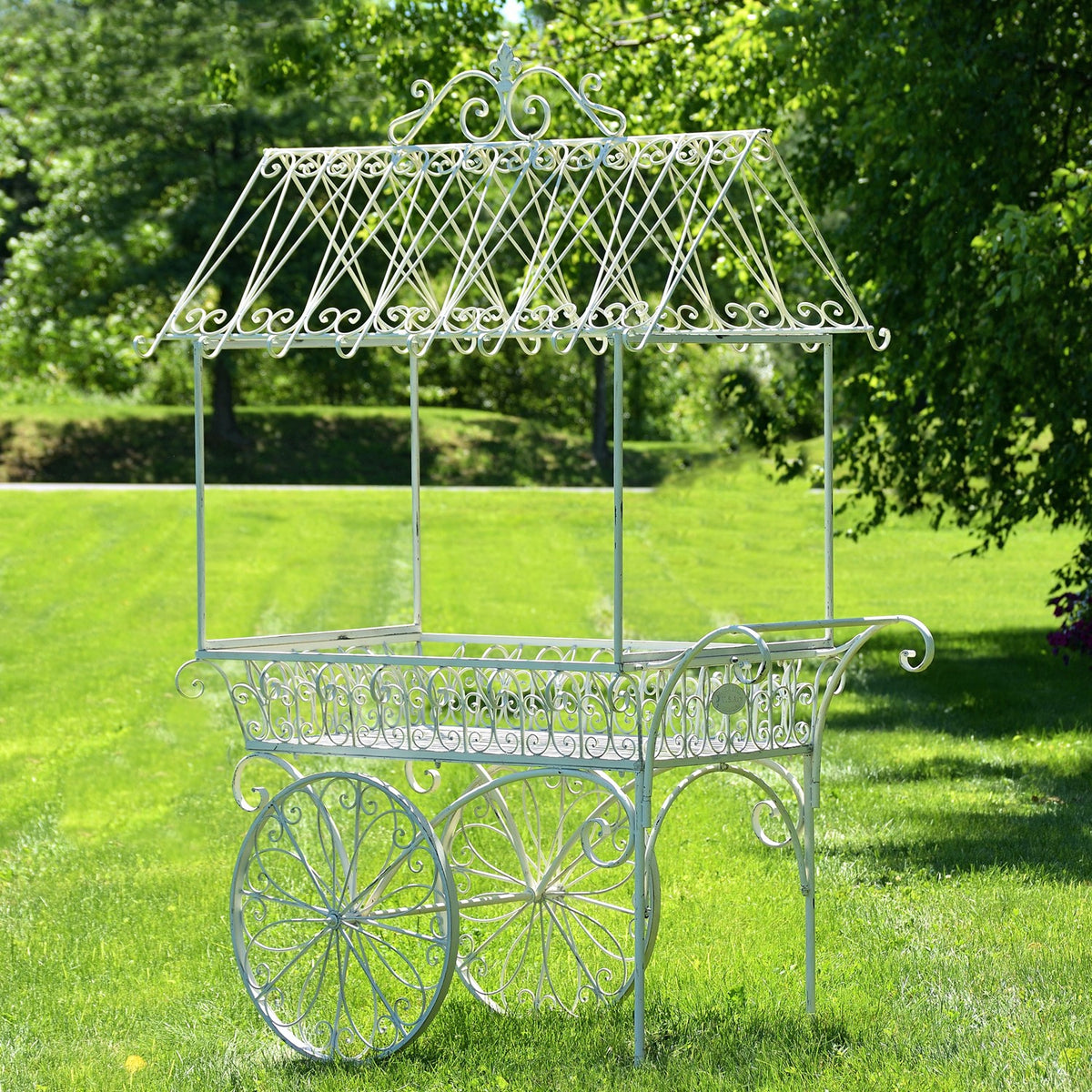 Empty white iron flower cart with an ornate peaked canopy and scrollwork, standing on grass.