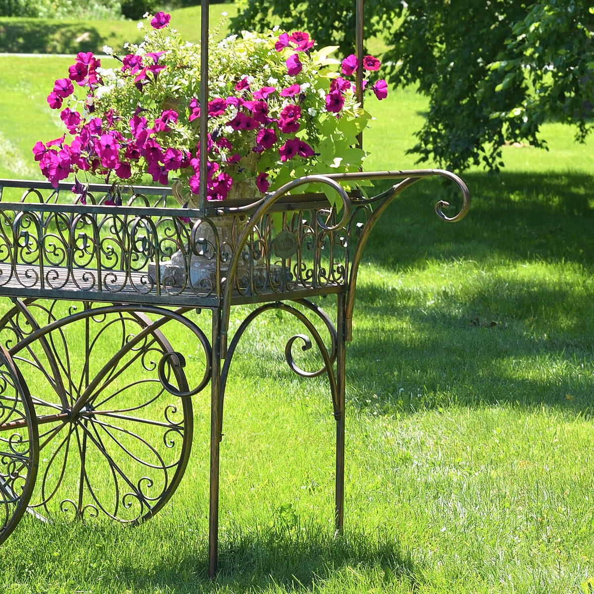 Magenta flowers fill the iron cart beside a curled handle and slender support legs.