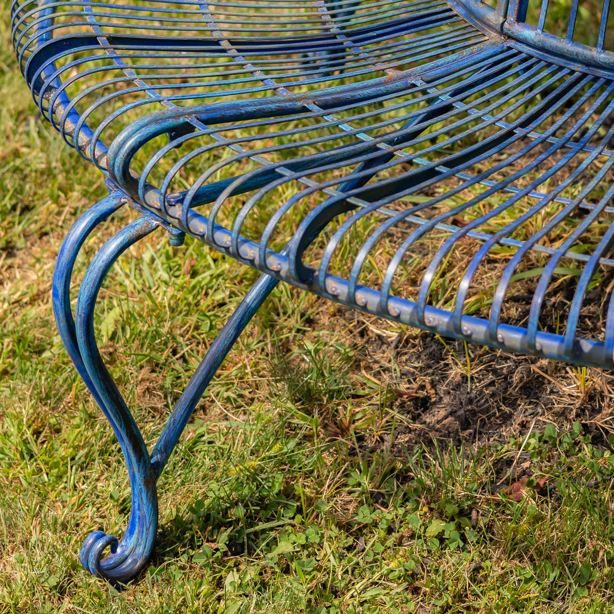 Close-up of the blue bench’s curved metal leg, curled foot, and radiating seat bars above grass.