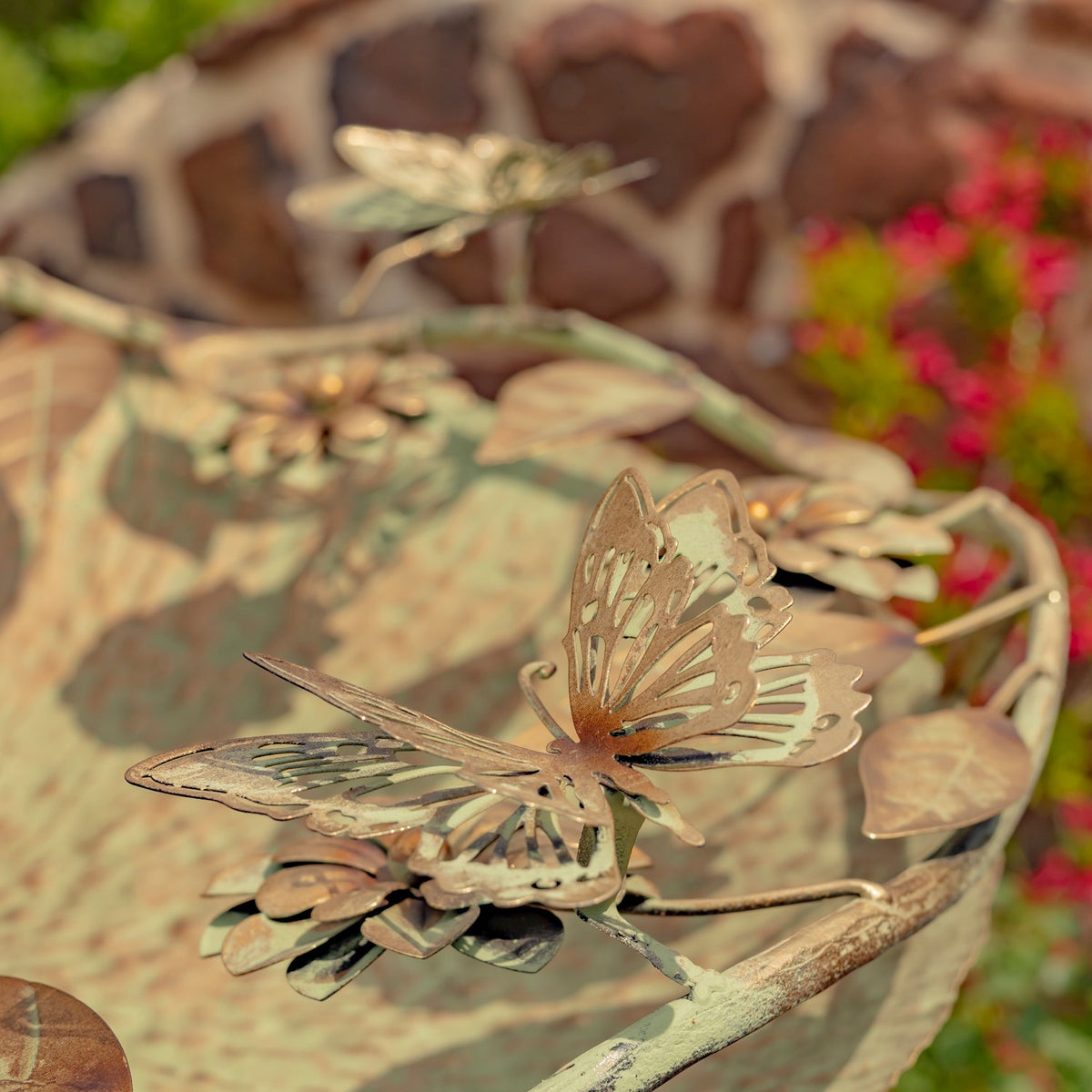 Metal butterfly ornament with openwork wings perched on a flower at the birdbath rim.