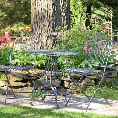 The bistro set is shown from the side in a garden, with a stone wall and tree behind it.
