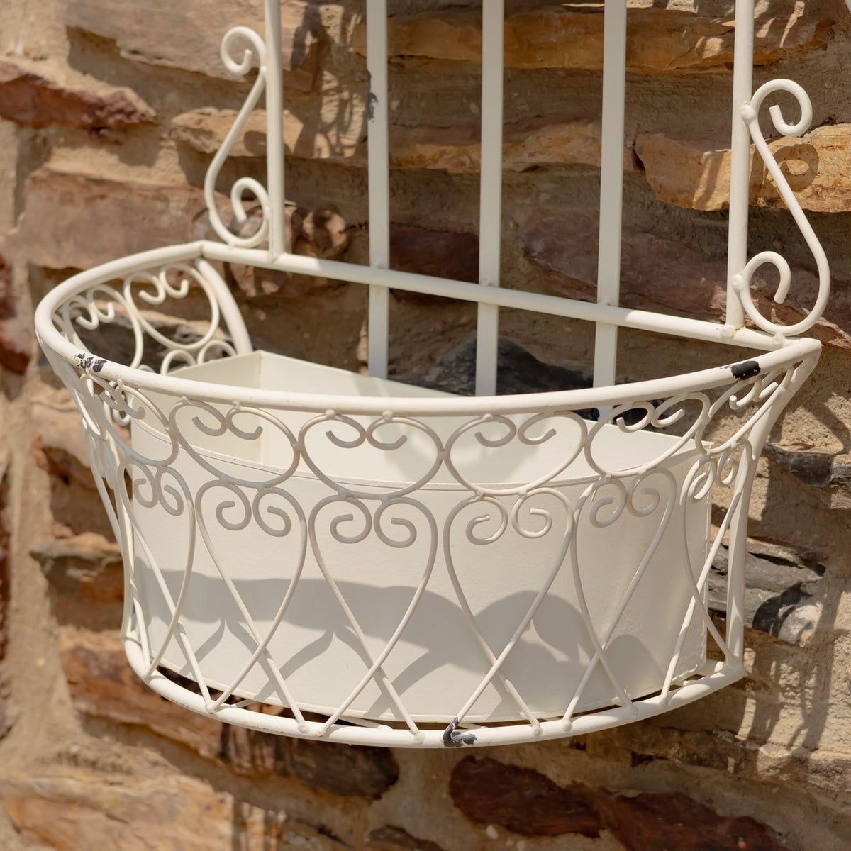 Antique-white planter basket with ornate curling metalwork hanging against a stone wall.