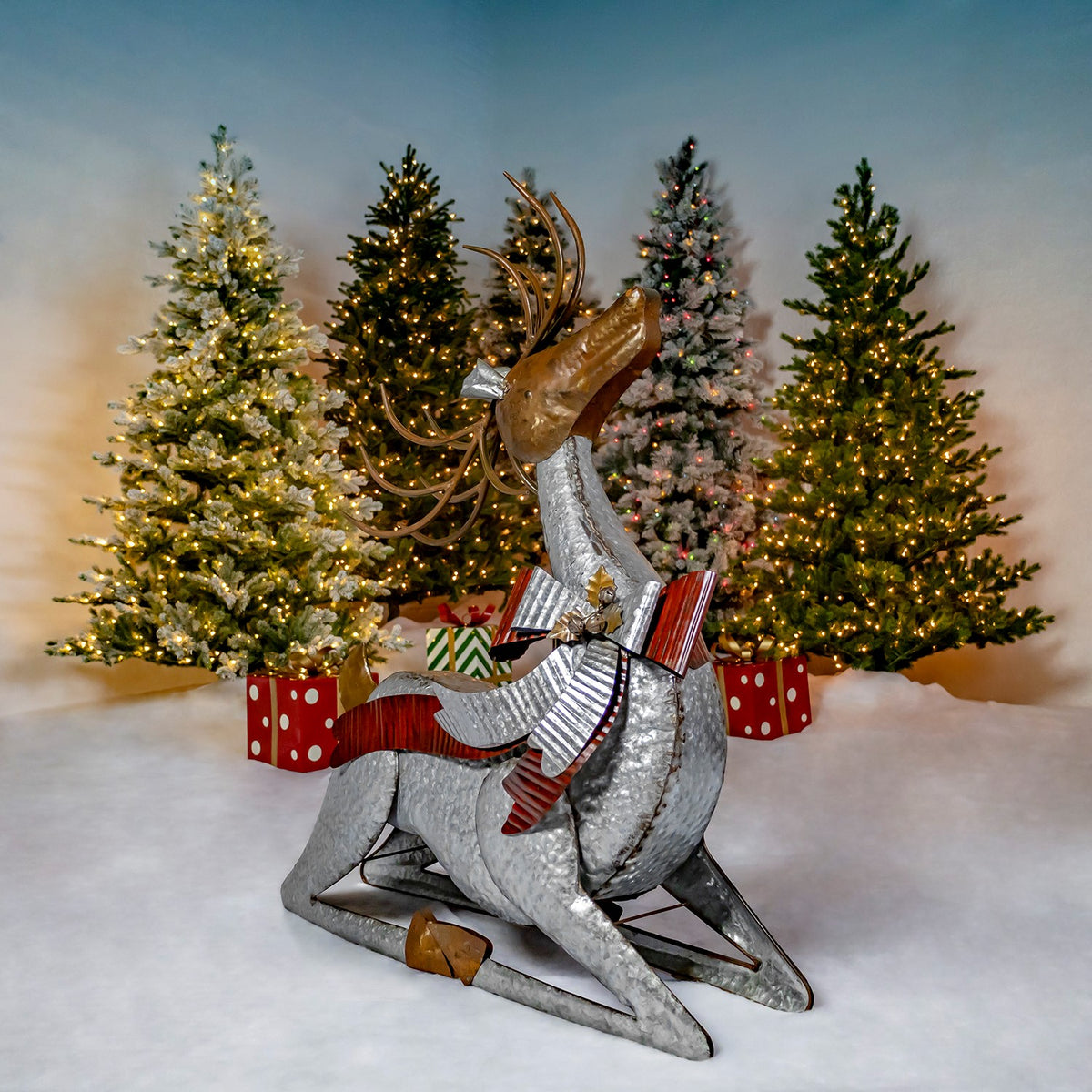 A silver reindeer rests on bent legs, with antlers, a striped bow and bell against a festive tree backdrop.