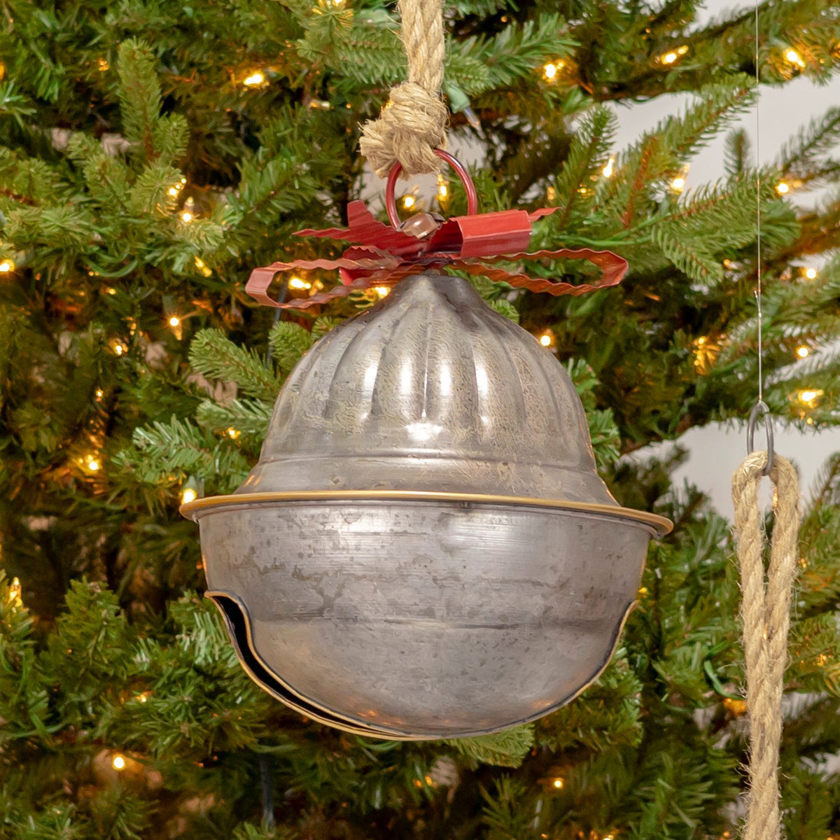 Close view of a rounded galvanized bell with a red bow and thick rope hanging against evergreen branches.