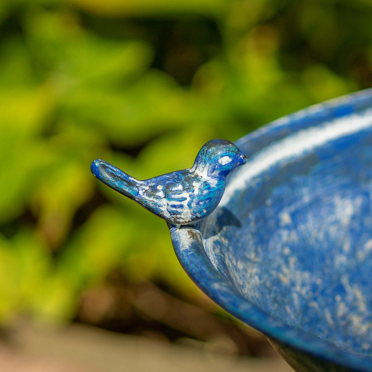 Close-up of a blue bird ornament perched on the curved rim of the birdbath.