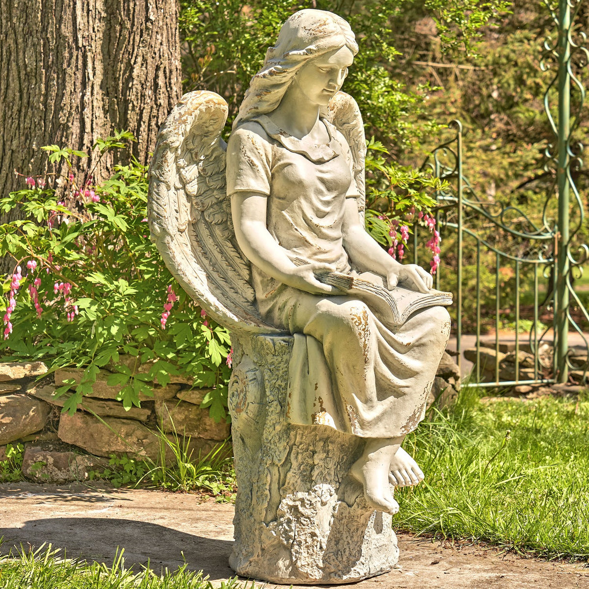 A seated angel statue looks down at an open book, shown among garden plants and stonework.