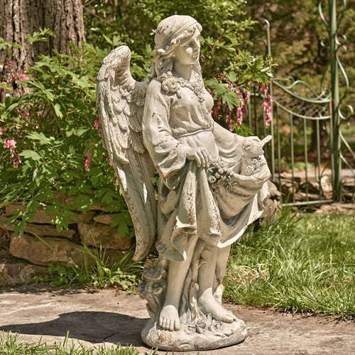 Winged angel statue holding a draped garment, standing among garden greenery.