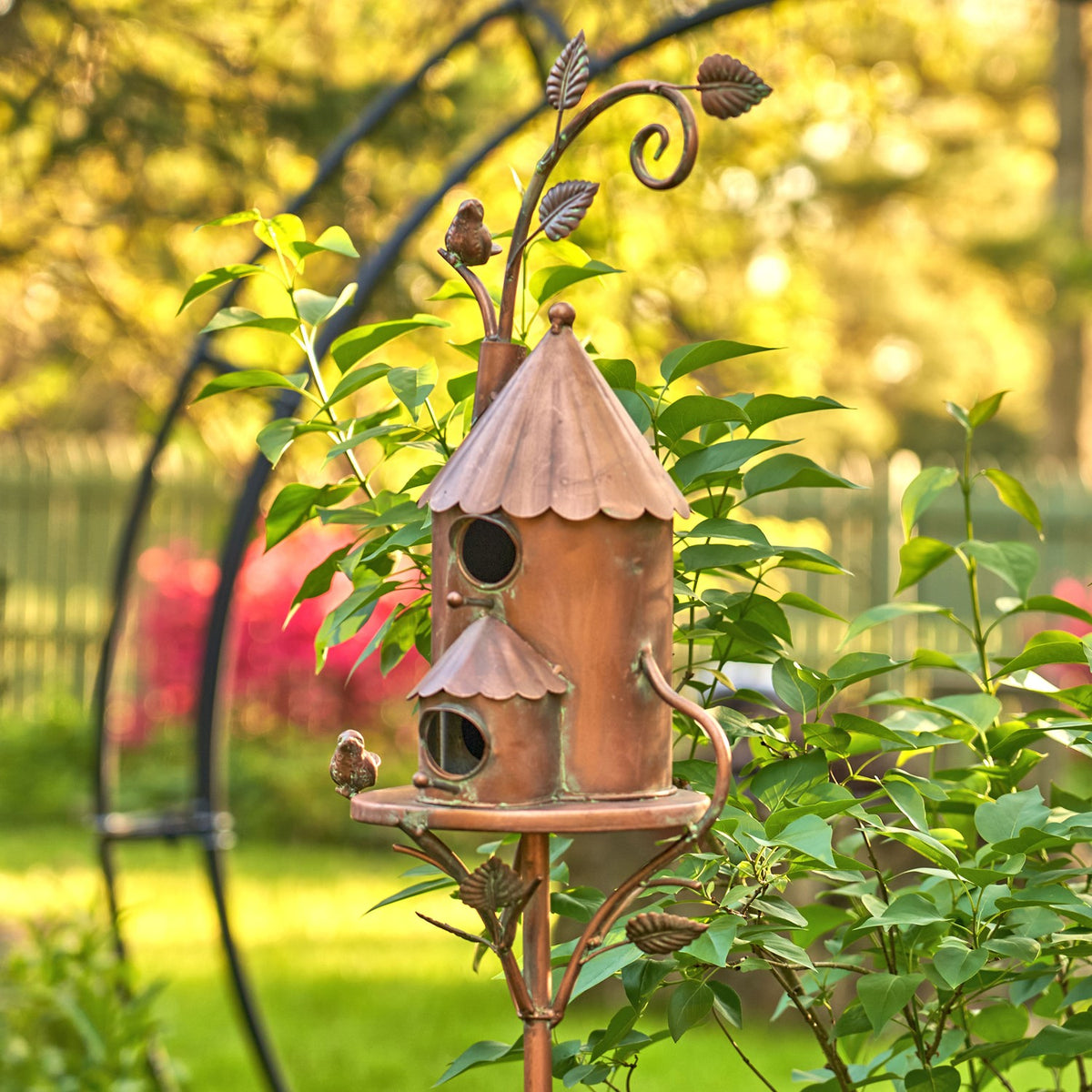 Two-story copper birdhouse stake viewed among garden shrubs from the opposite side