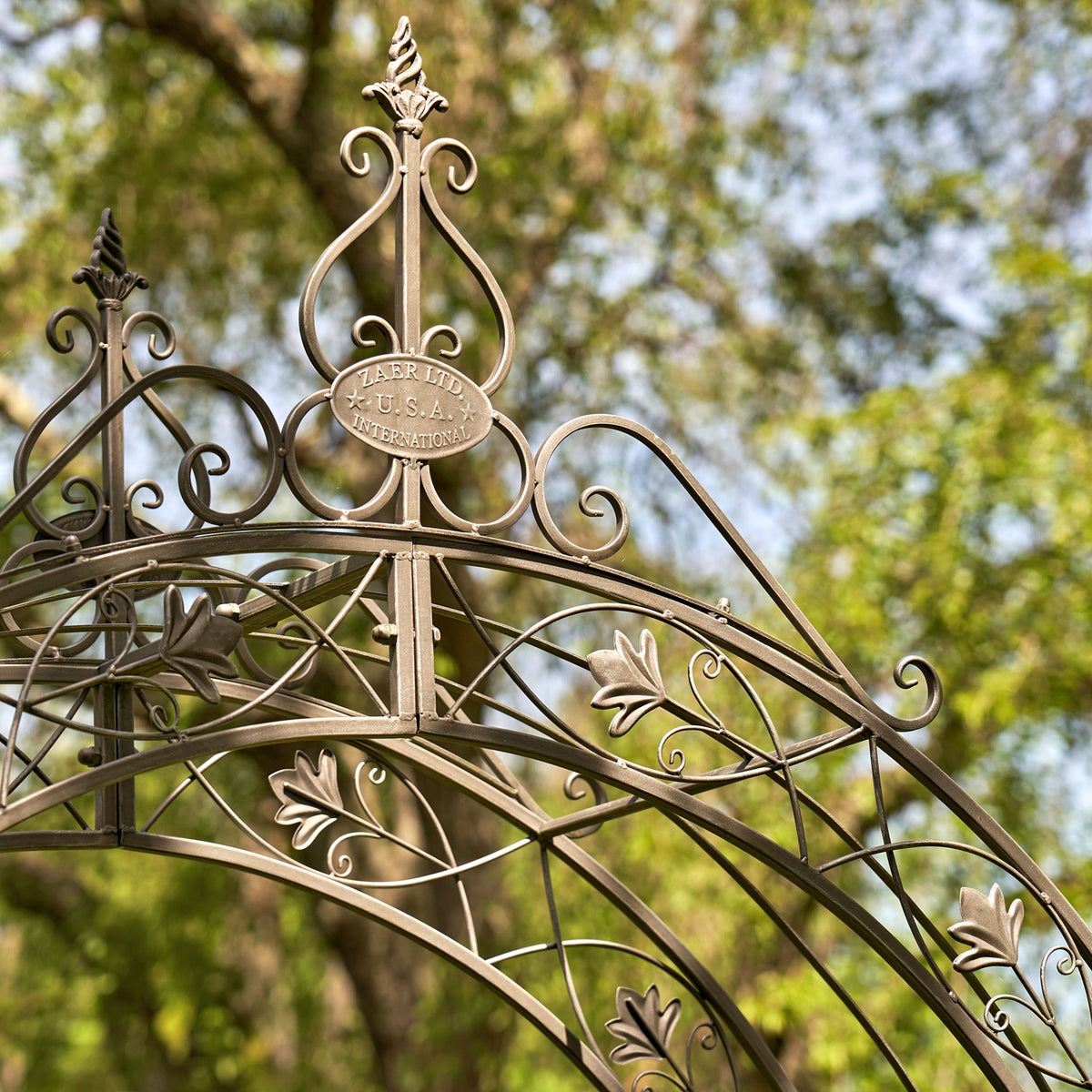 Close-up of the garden arch’s curved ironwork, leaf accents and central oval plaque against leafy trees.