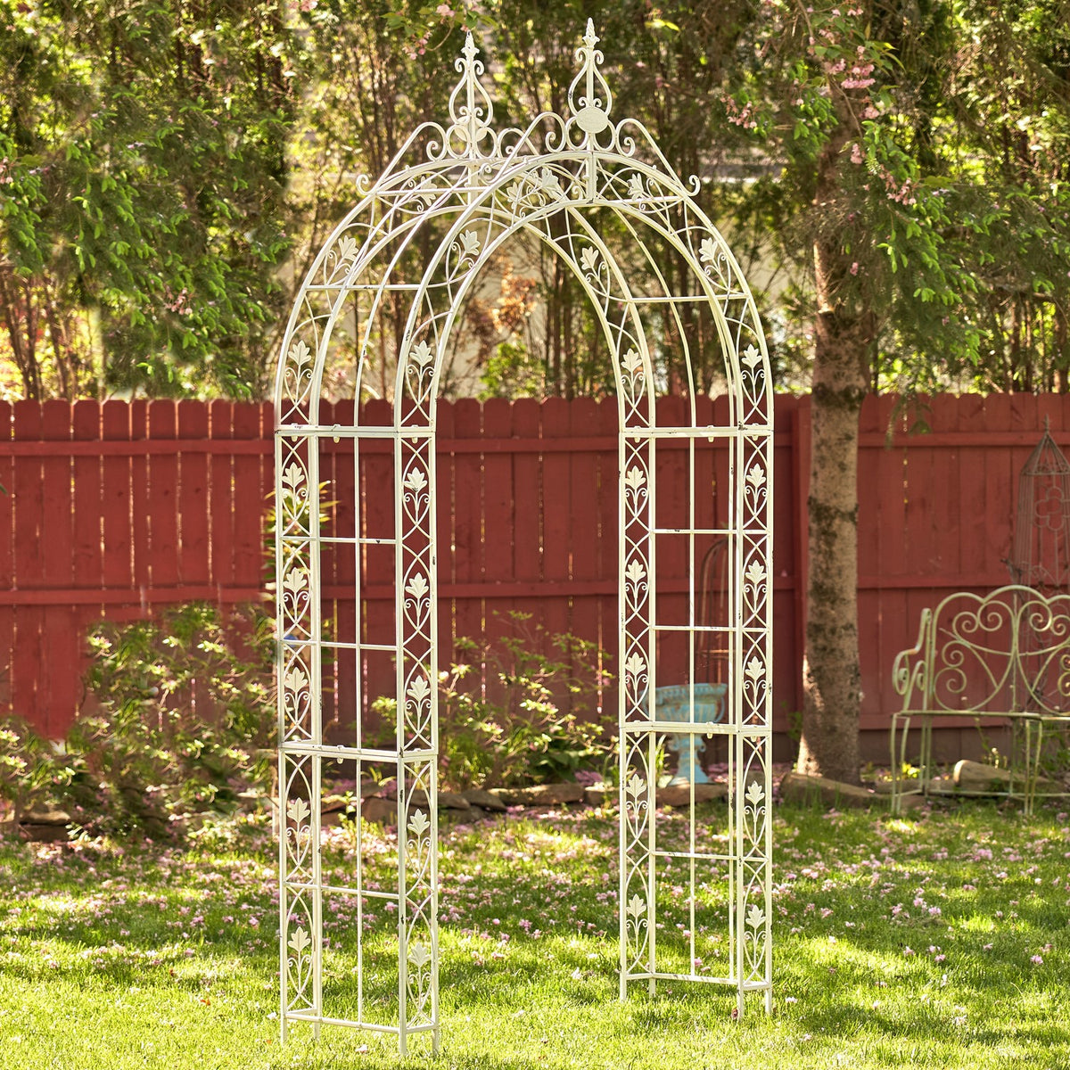 Pale metal garden arch shown on grass in front of a red wooden fence.