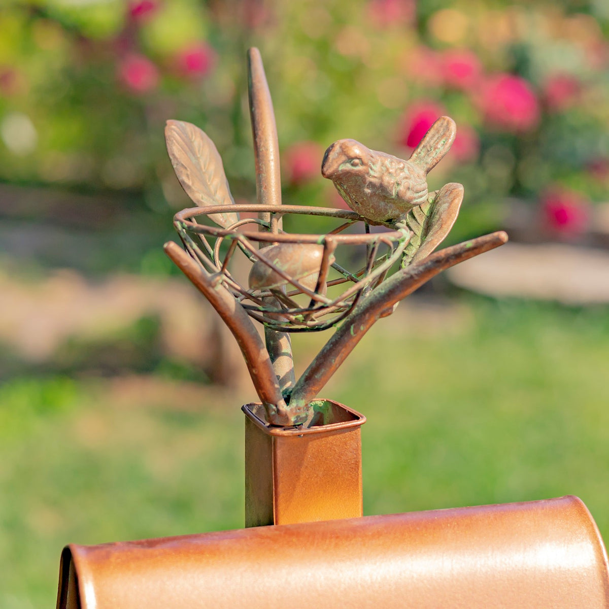 Close-up of a small metal bird perched beside a twig nest ornament above the birdhouse roof.