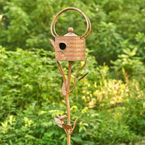 Teapot birdhouse and decorative stake shown against leafy garden greenery