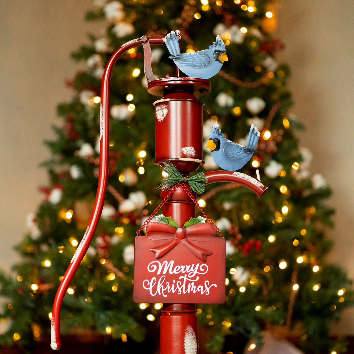 Red water pump ornament with two blue birds, a bow, greenery, and a hanging red holiday sign.