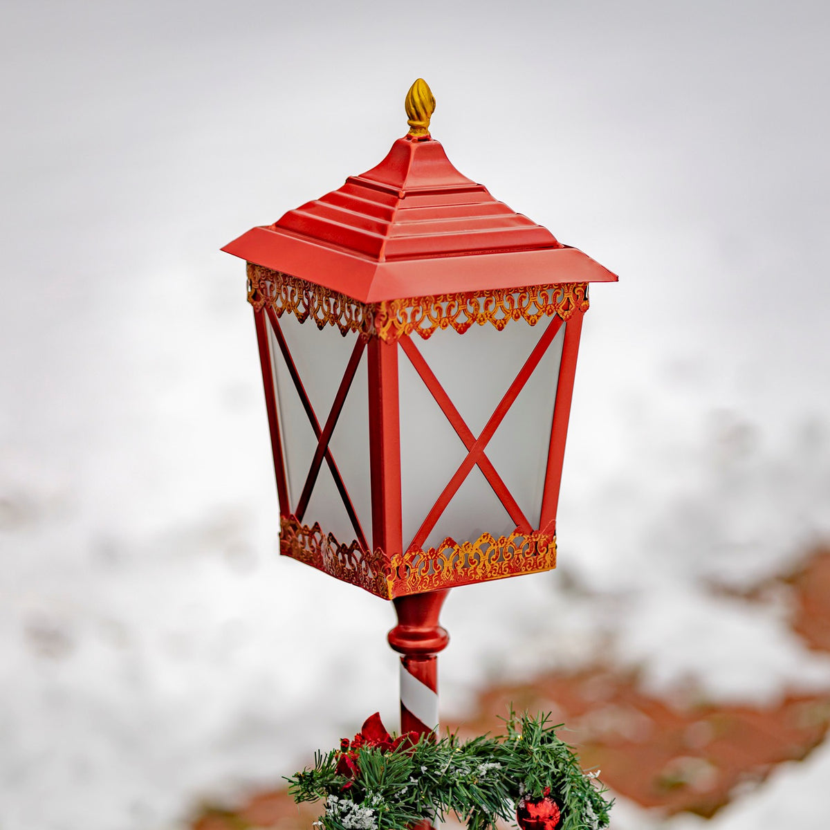 Red lantern with a peaked roof, crisscross supports, and ornate trim above a greenery wreath.
