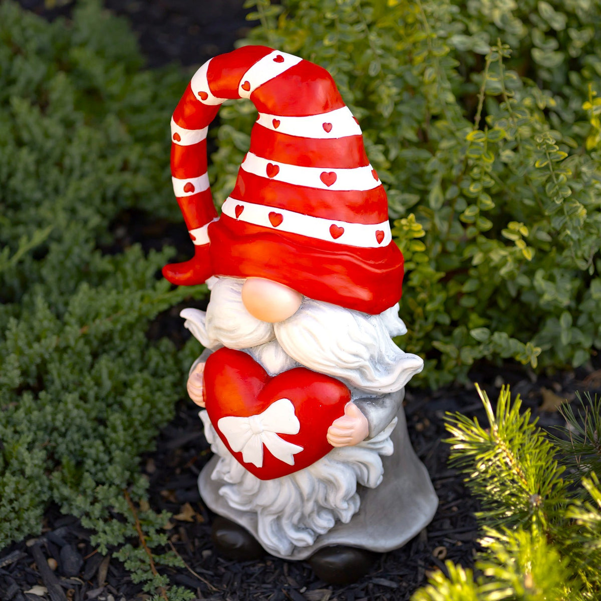 Valentine gnome holding a red heart, angled garden view with green foliage