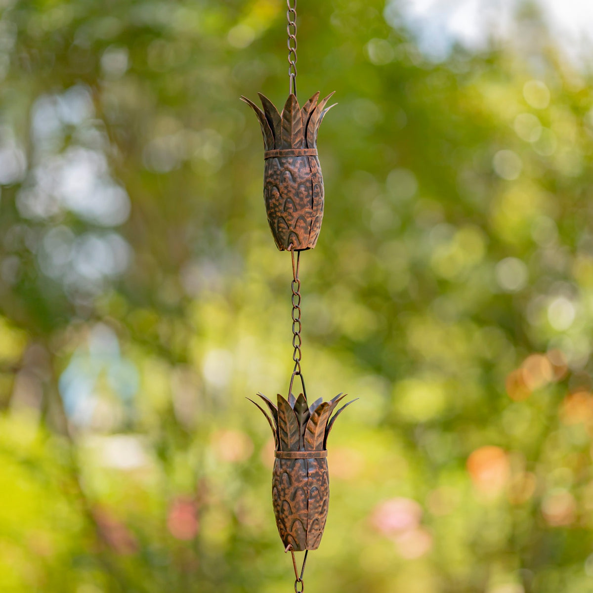 Two antique bronze pineapple-shaped links on a rain chain against a leafy garden
