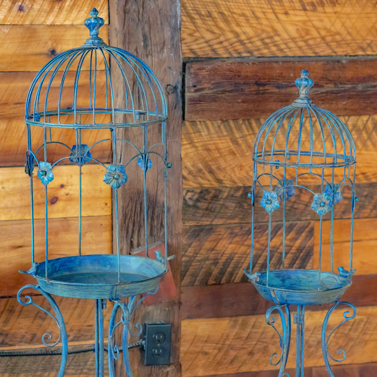 Two blue iron birdcage planters with flower-shaped accents and shallow trays against a wood plank wall.