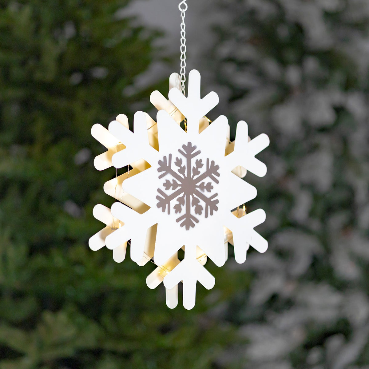 White hanging snowflake with silver center shown against a blurred evergreen tree