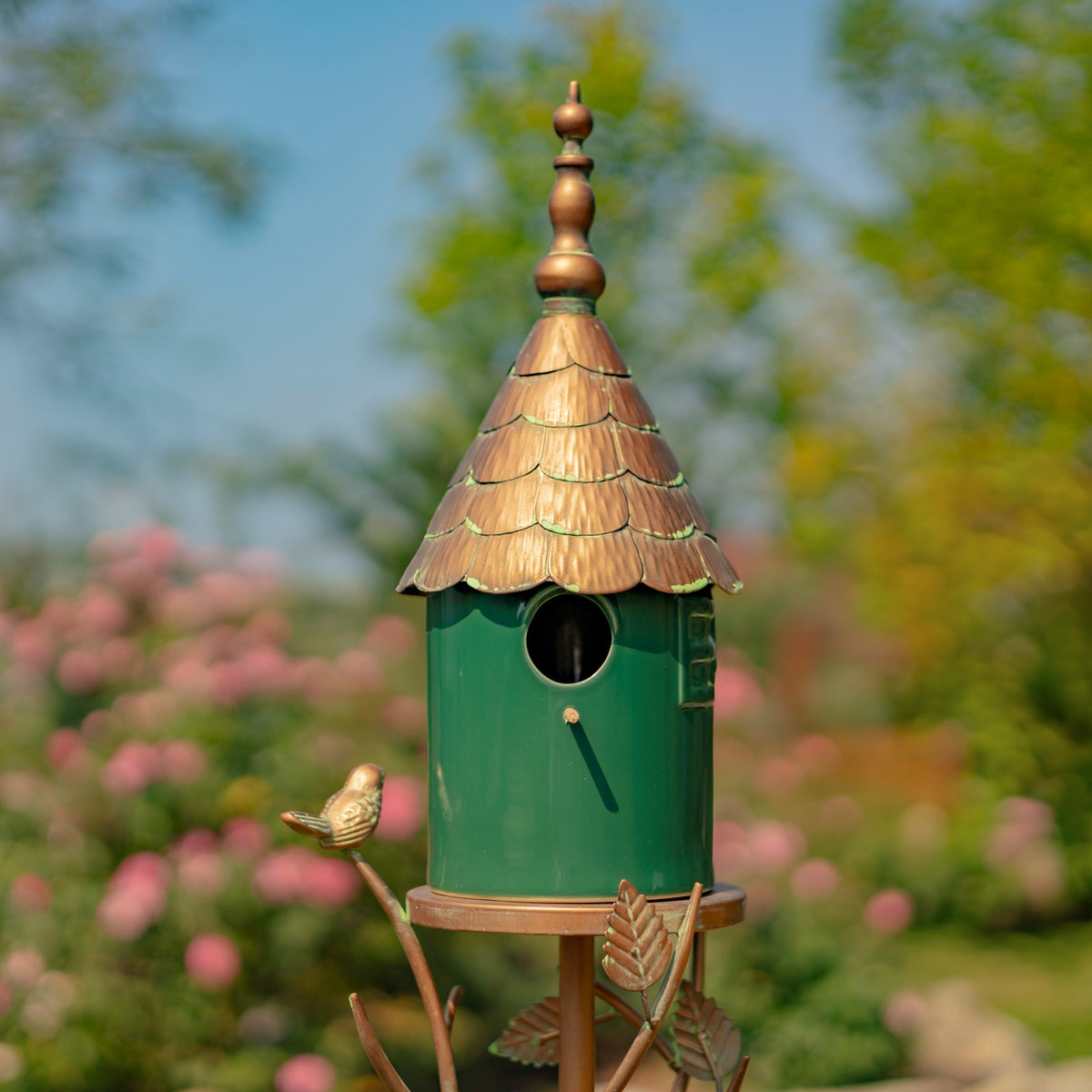Closer view of the green birdhouse, round entrance, layered copper-colored roof and small metal bird ornament.