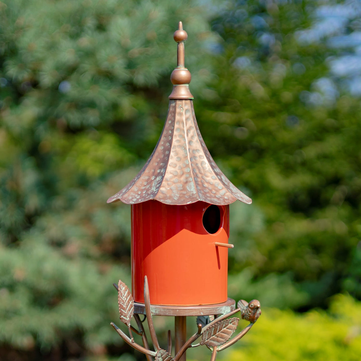 Close view of the red birdhouse, showing its round entrance, small perch, and scalloped metal roof.