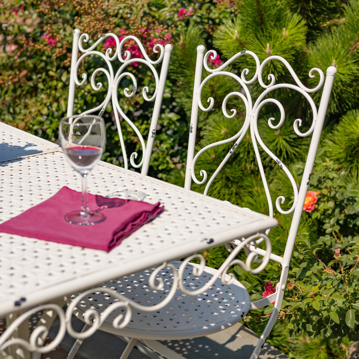 Antique white metal table and two matching chairs with ornate scrollwork on a garden patio.