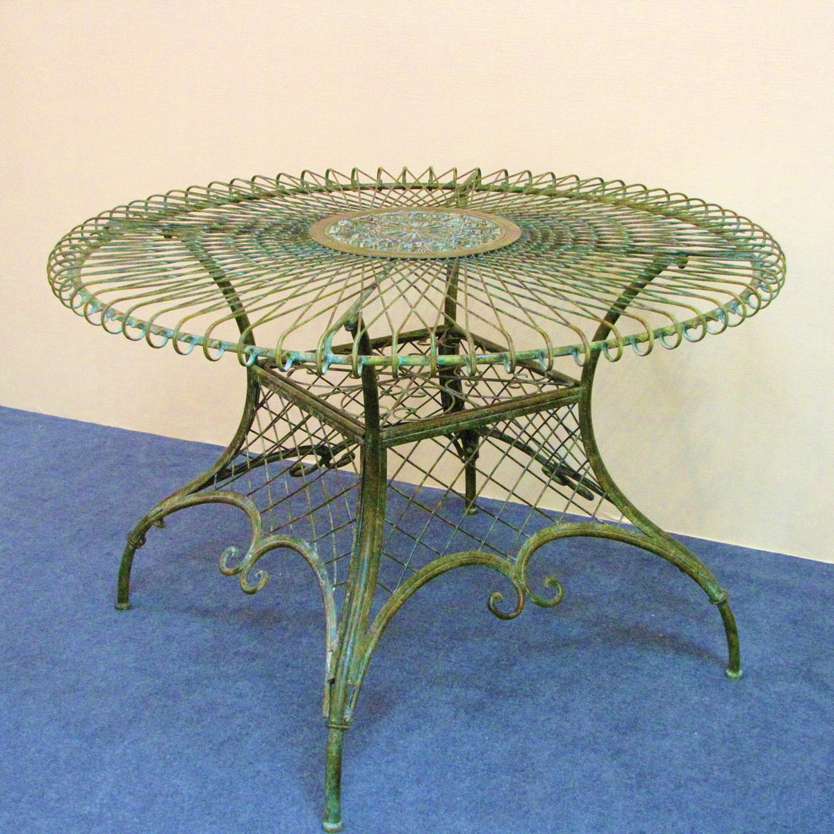 Round green iron table with an open lattice top and decorative curved supports against a plain wall.
