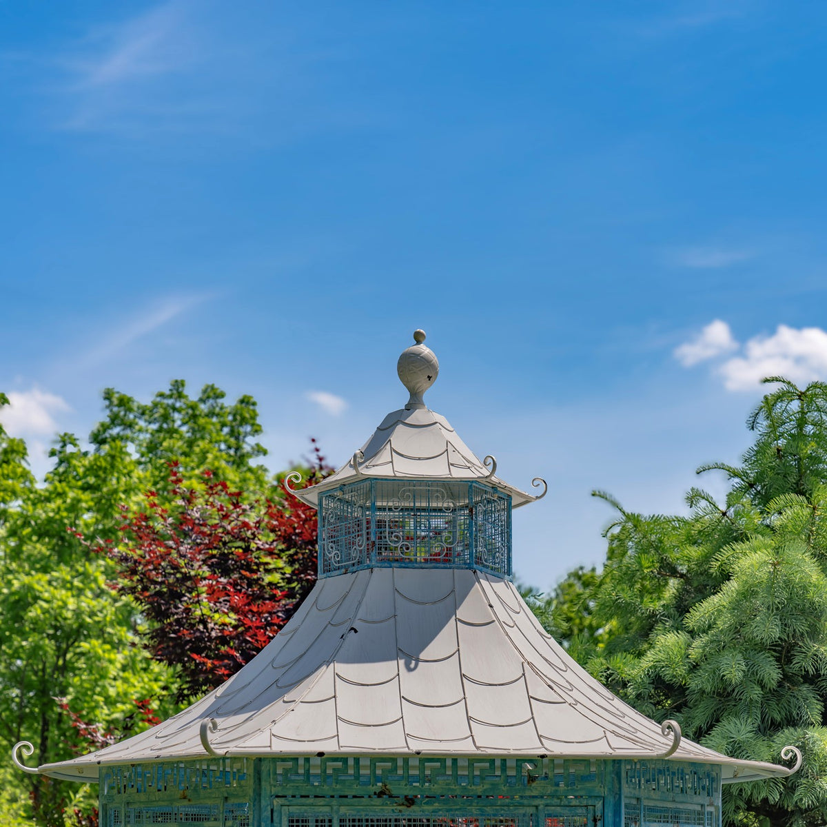 Close view of the gazebo’s tiered pagoda roof, ornamental finial, and decorative metalwork against a blue sky.