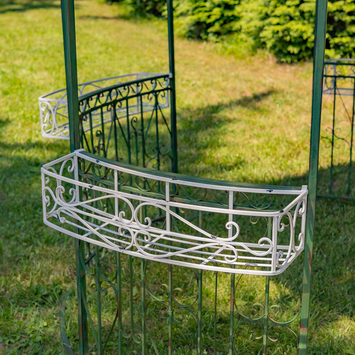 Curved pale metal basket rail with scrollwork attached to the gazebo’s green frame.