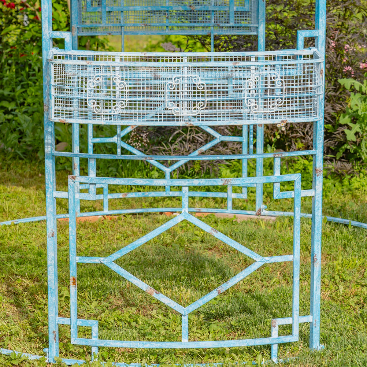 Close view of a curved metal planter and geometric iron panels, with the gazebo's blue finish visibly weathered.