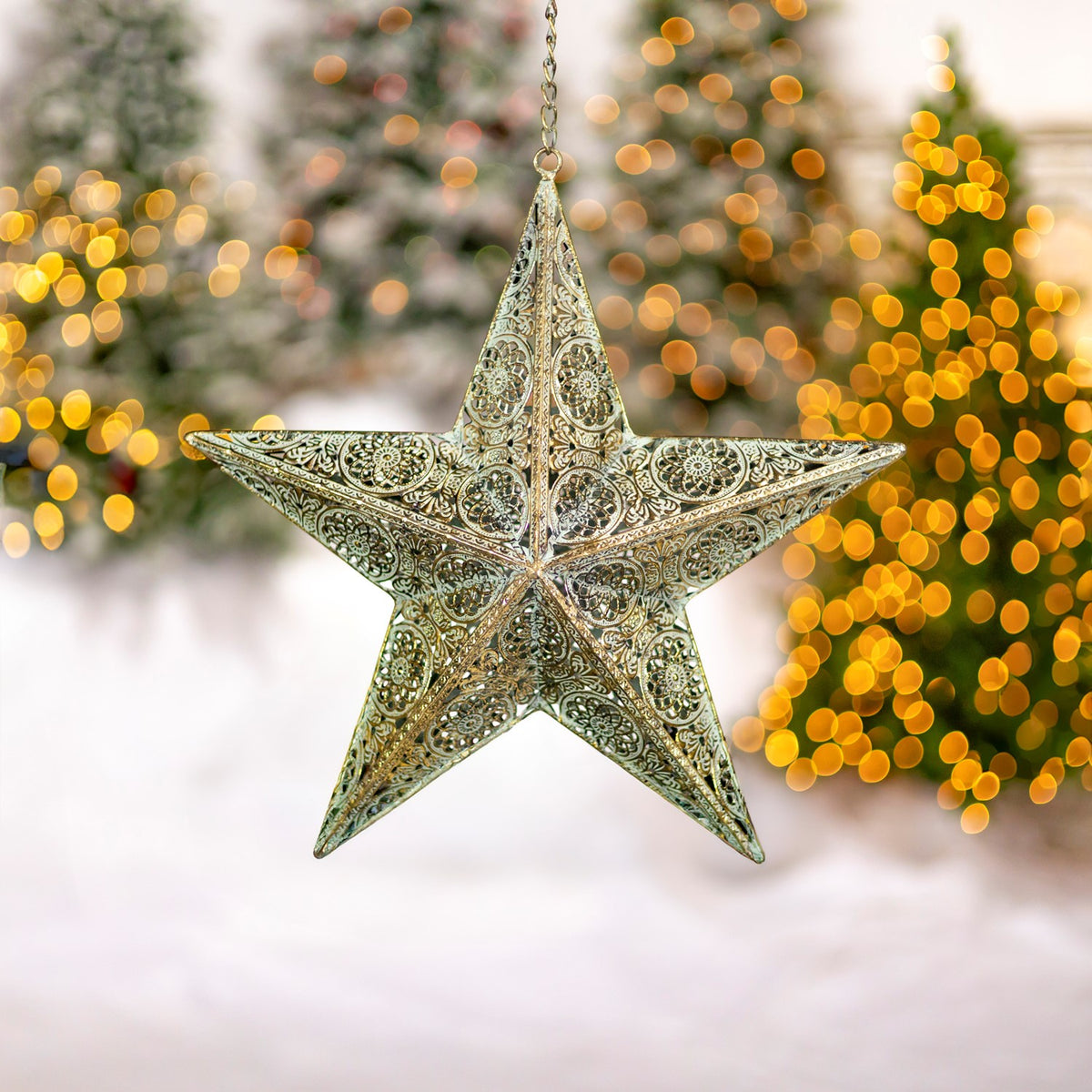 A hanging gold star ornament with raised points and intricate cutout patterns is shown against blurred tree lights.