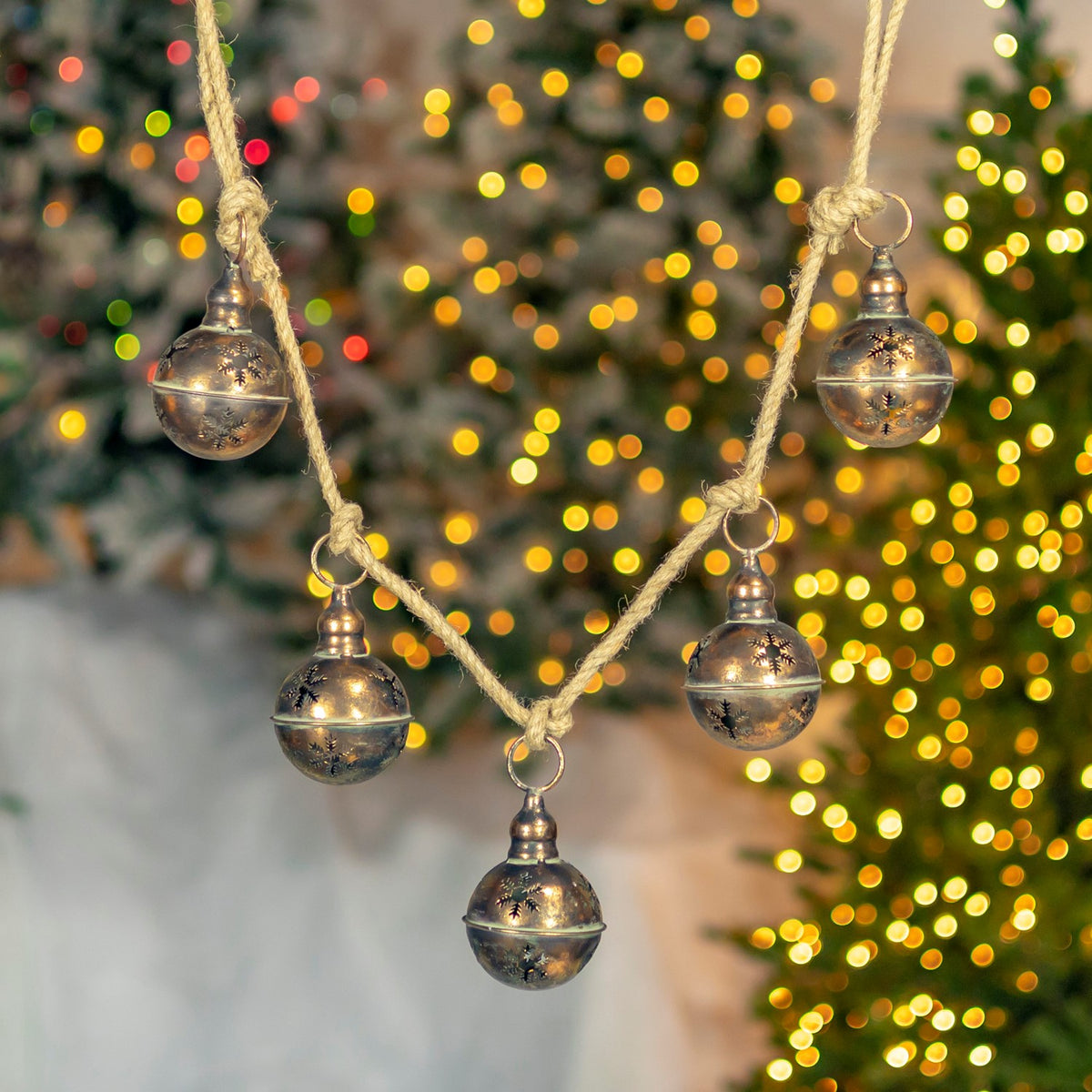 Five round antique-gold bells hang from a rope, with a decorated Christmas tree blurred behind them.