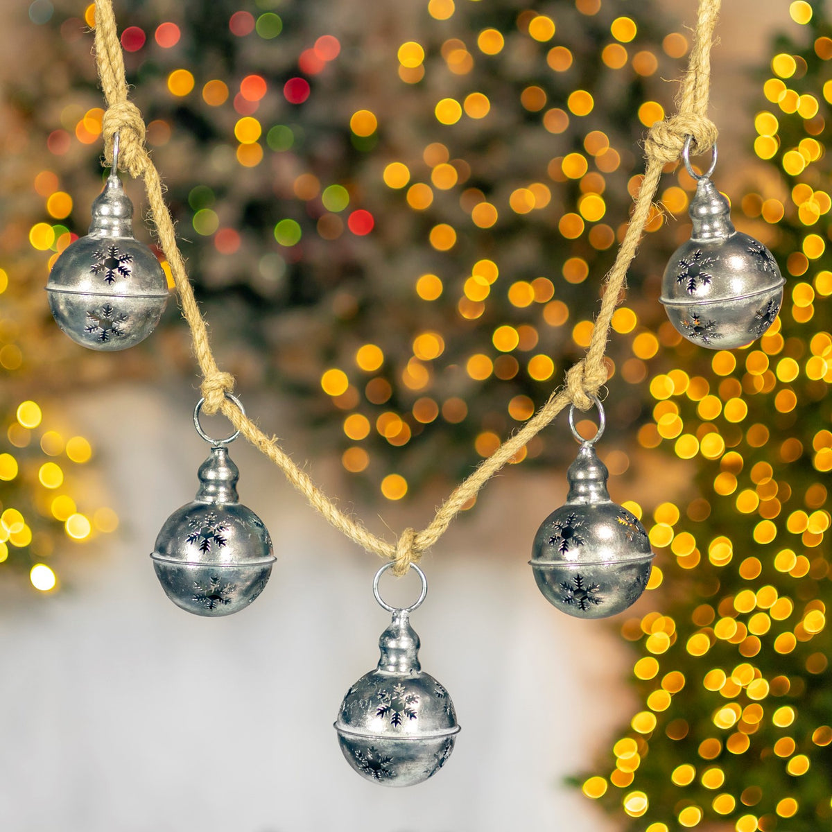 Five round silver bells with dark snowflake motifs dangle from twine against golden bokeh.