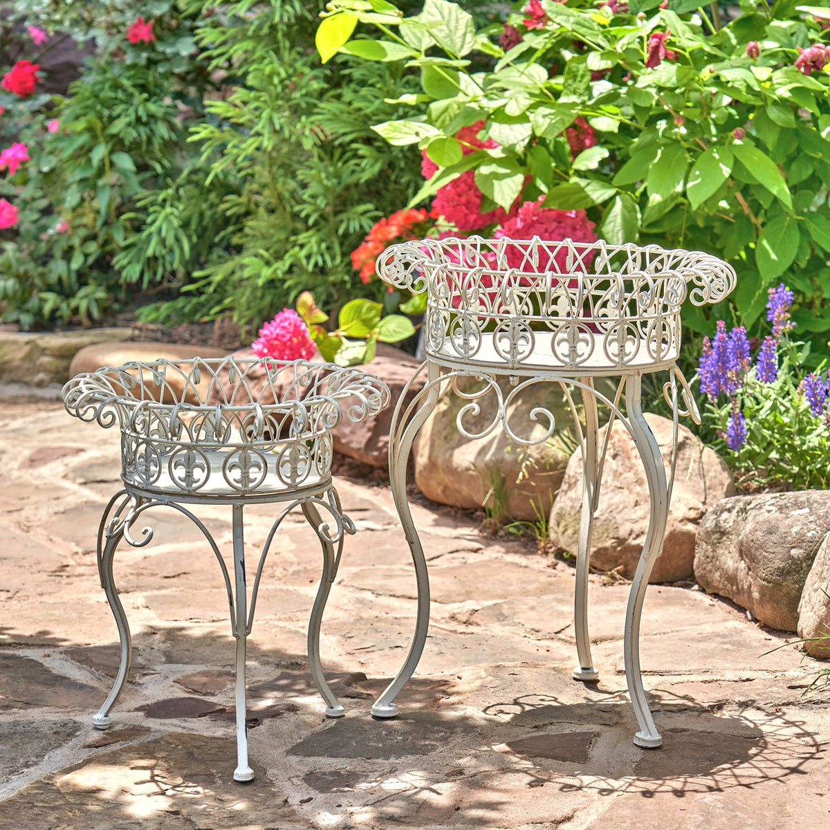 Two light-colored metal stands with round open baskets and curved legs beside garden flowers.