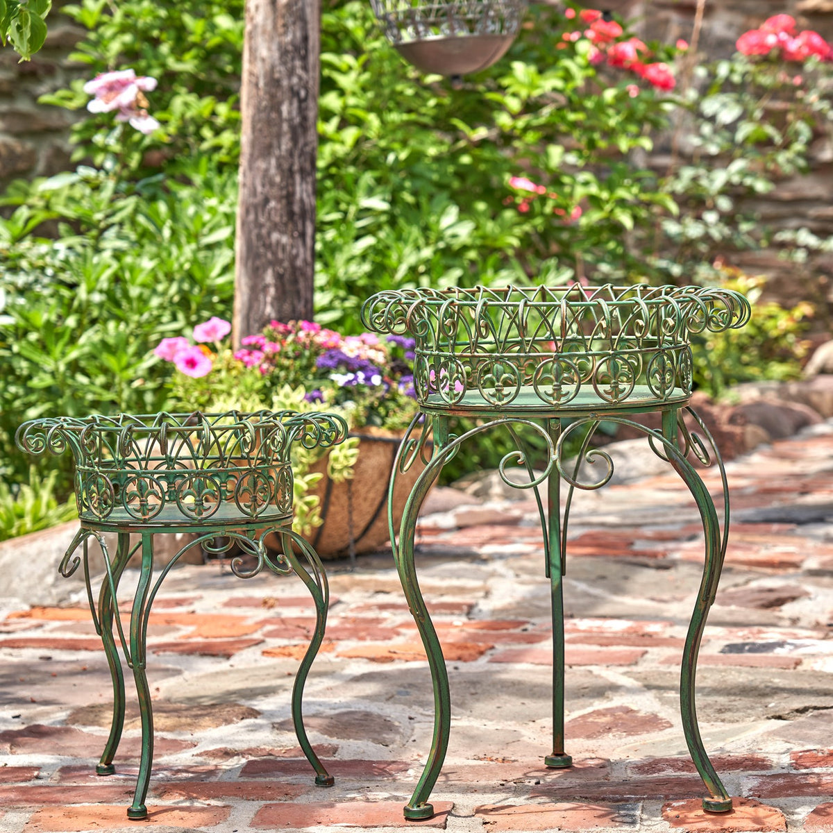 Two green metal plant stands of different heights displayed on a garden path among plants and flowers.