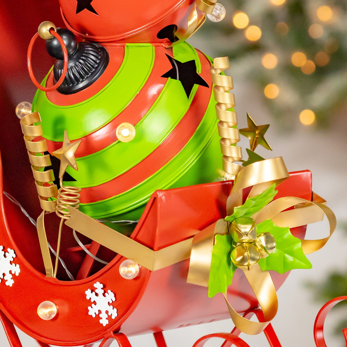 Close-up of the sleigh’s gold ribbon and bell cluster beside the green snowman body and snowflake decorations.