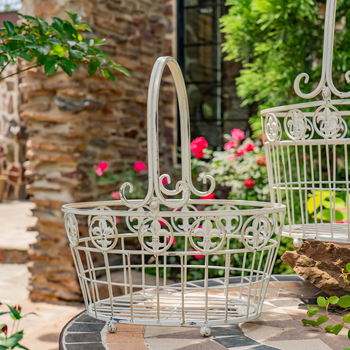Ornate pale metal basket with a tall handle on a tiled patio, with another basket partly visible behind it.