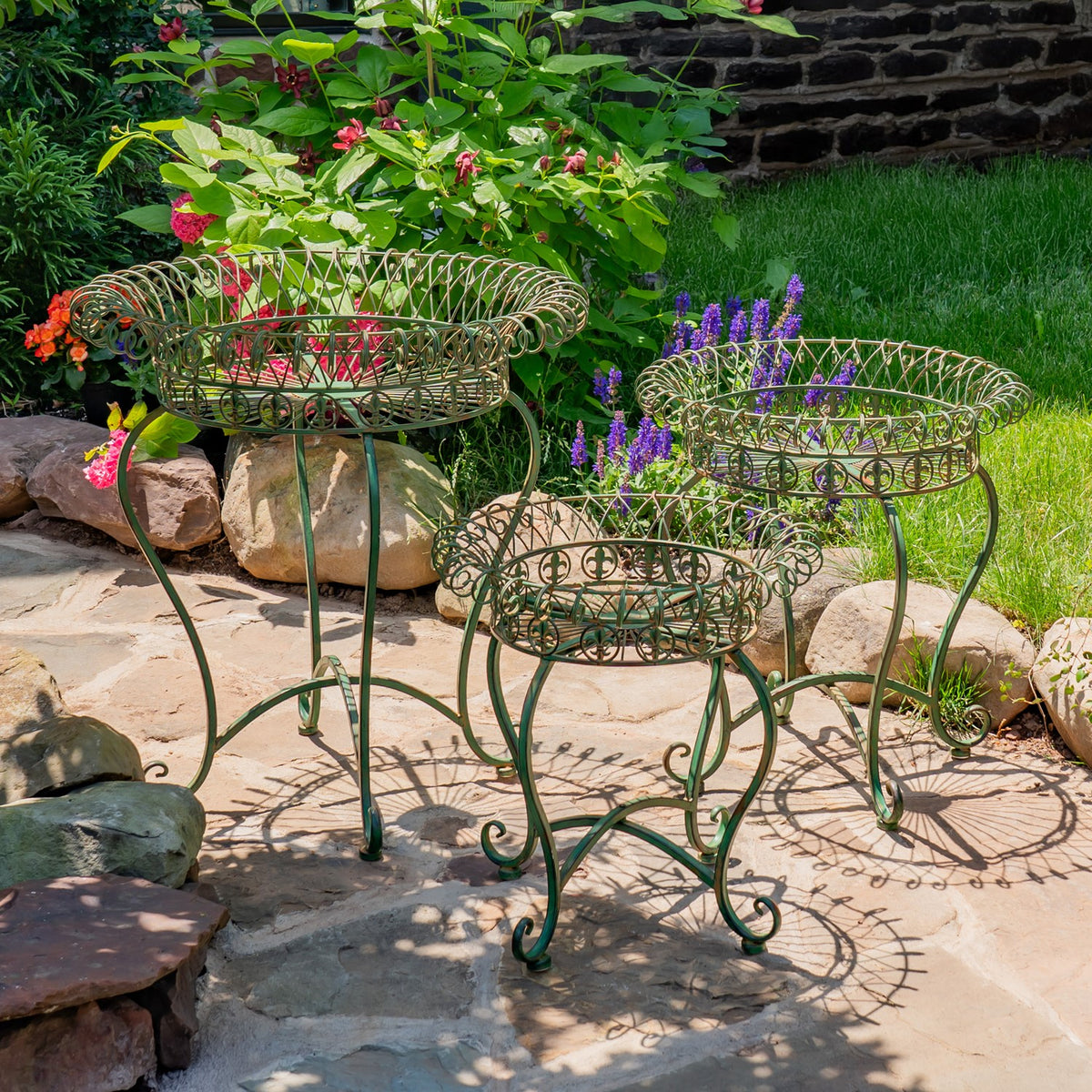 Three round wire plant stands on a stone patio, with leafy plants and flowers in two baskets.