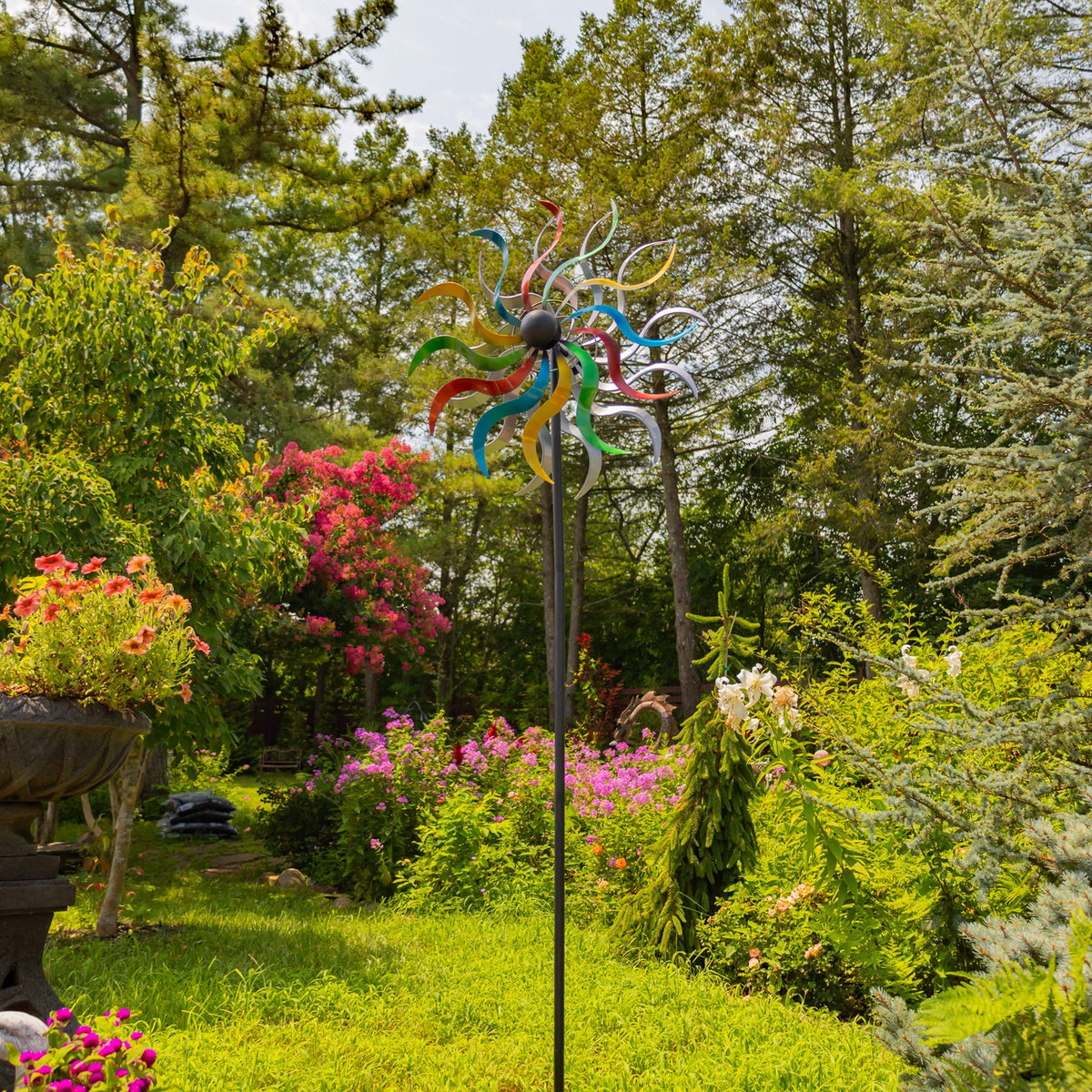 Tall wind spinner standing in a flower-filled garden, with its stake visible against the greenery.