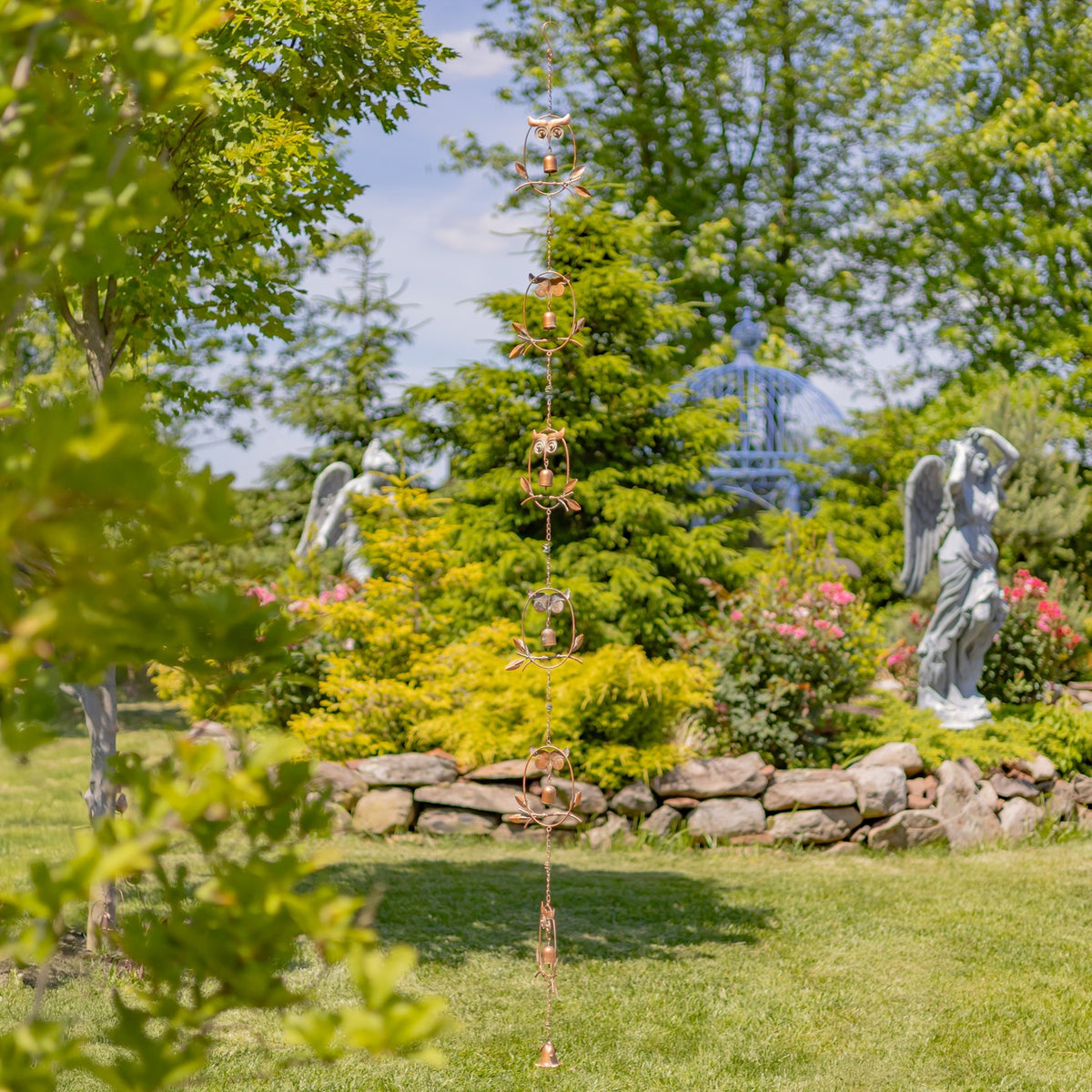 Long decorative rain chain hanging in a garden, with a stone wall and statues behind it.