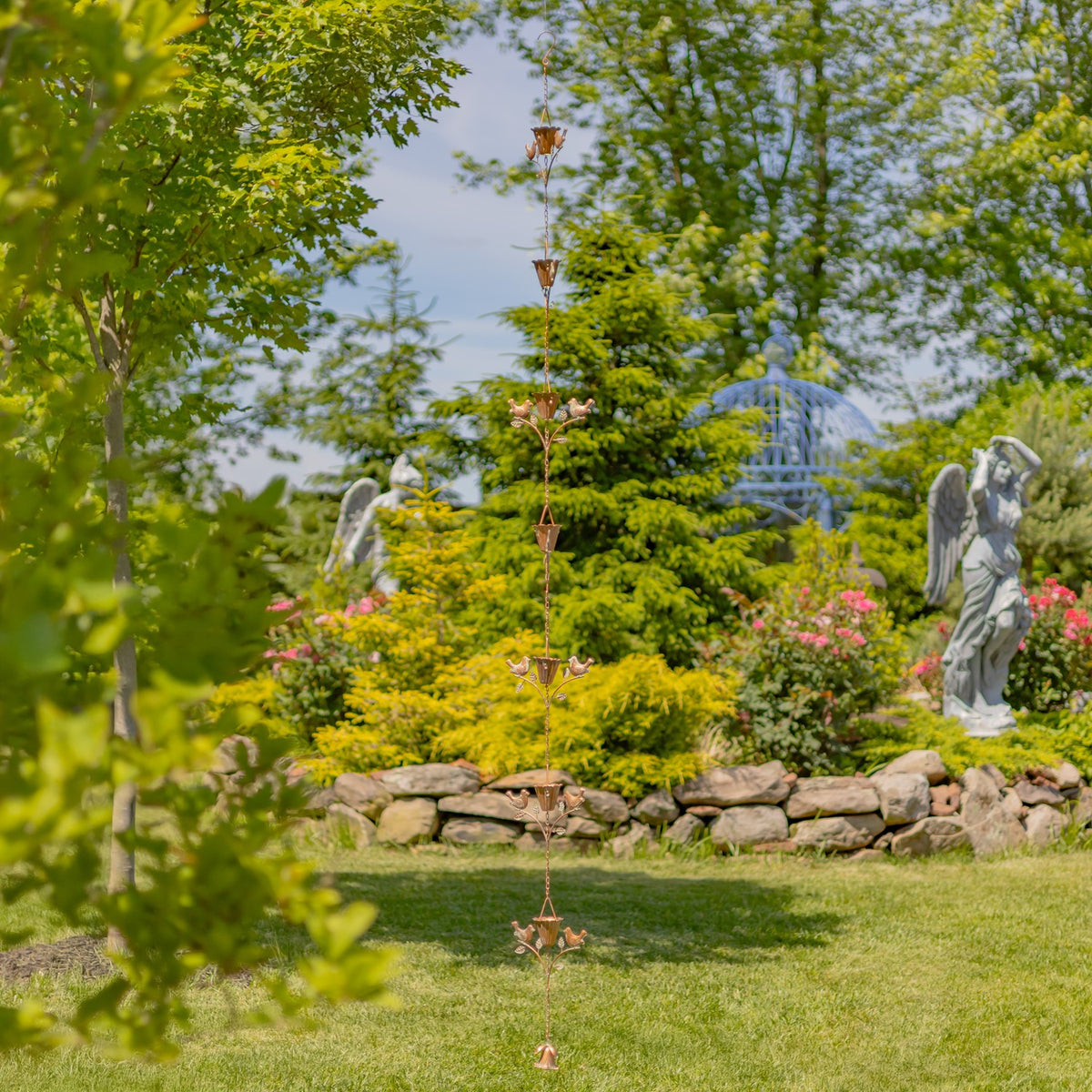 Rain chain hanging nearly to the lawn, framed by trees, stone edging, flowers, and garden statues.