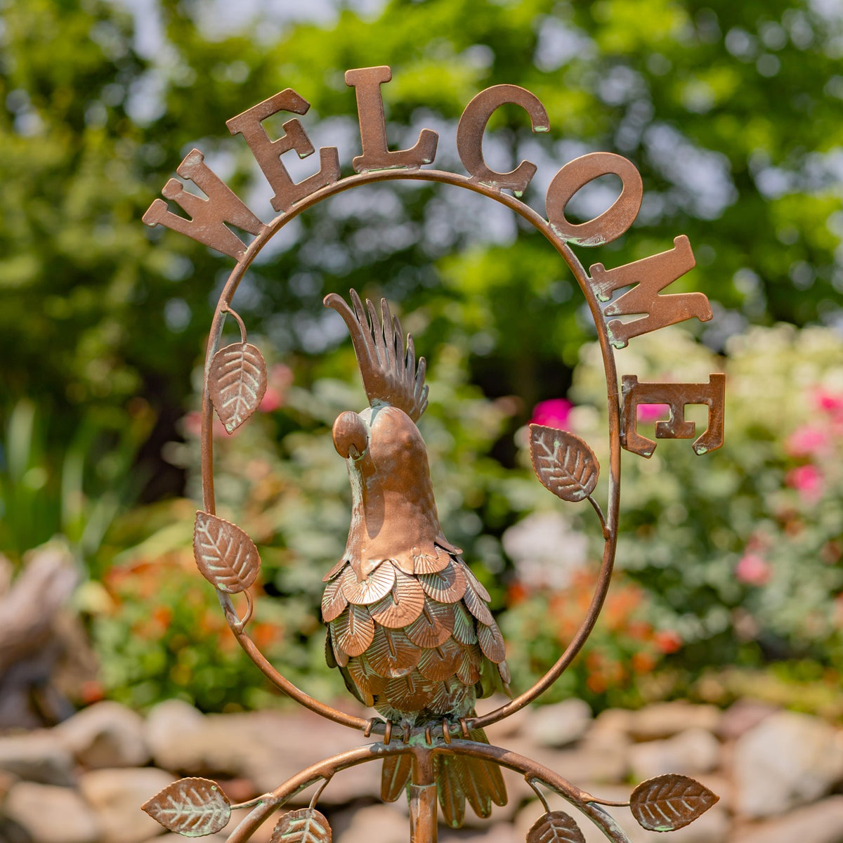 Closer view of the cockatoo ornament, layered metal feathers and leafy frame against a garden background.