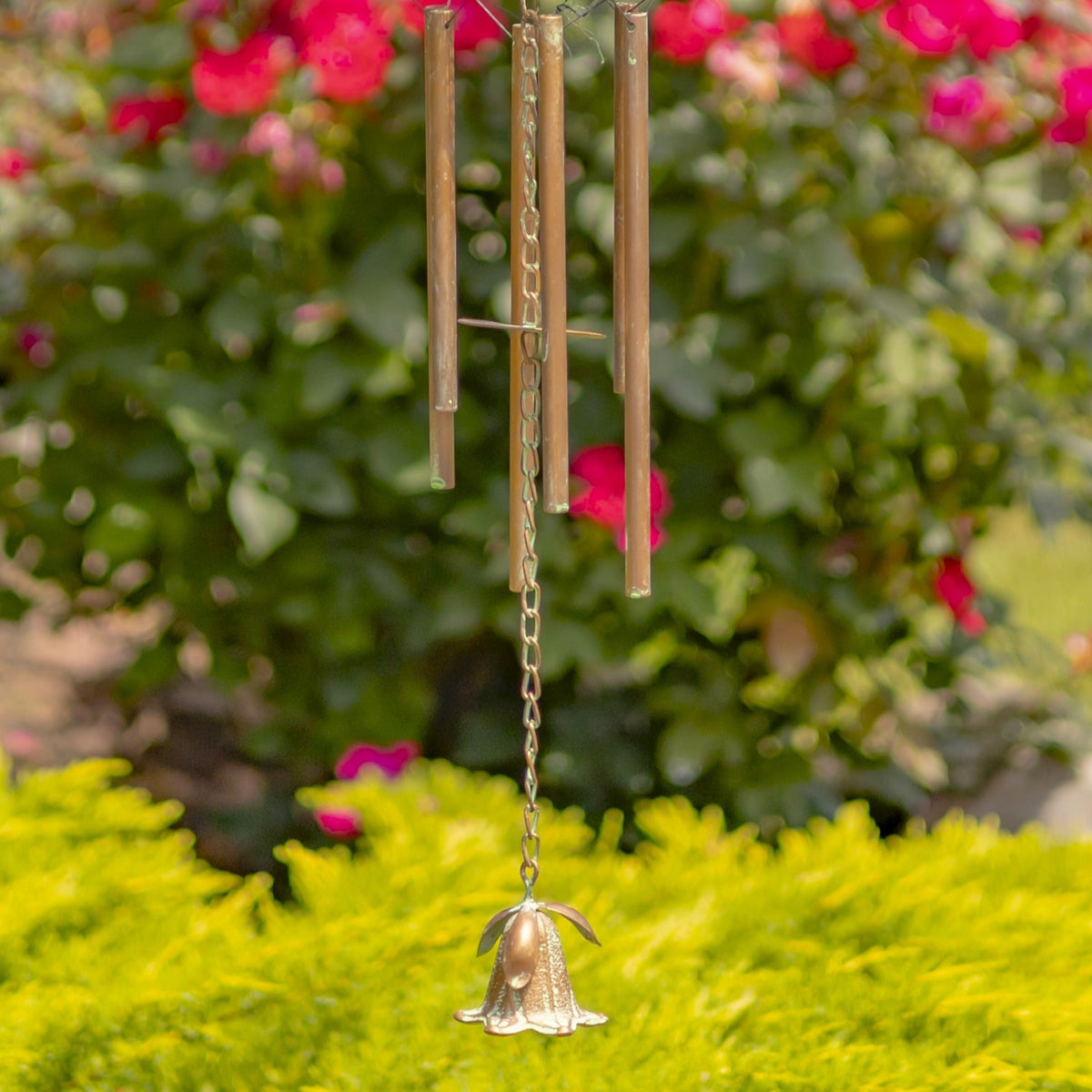 Long metal chime tubes and a small bell hang above bright green garden foliage.