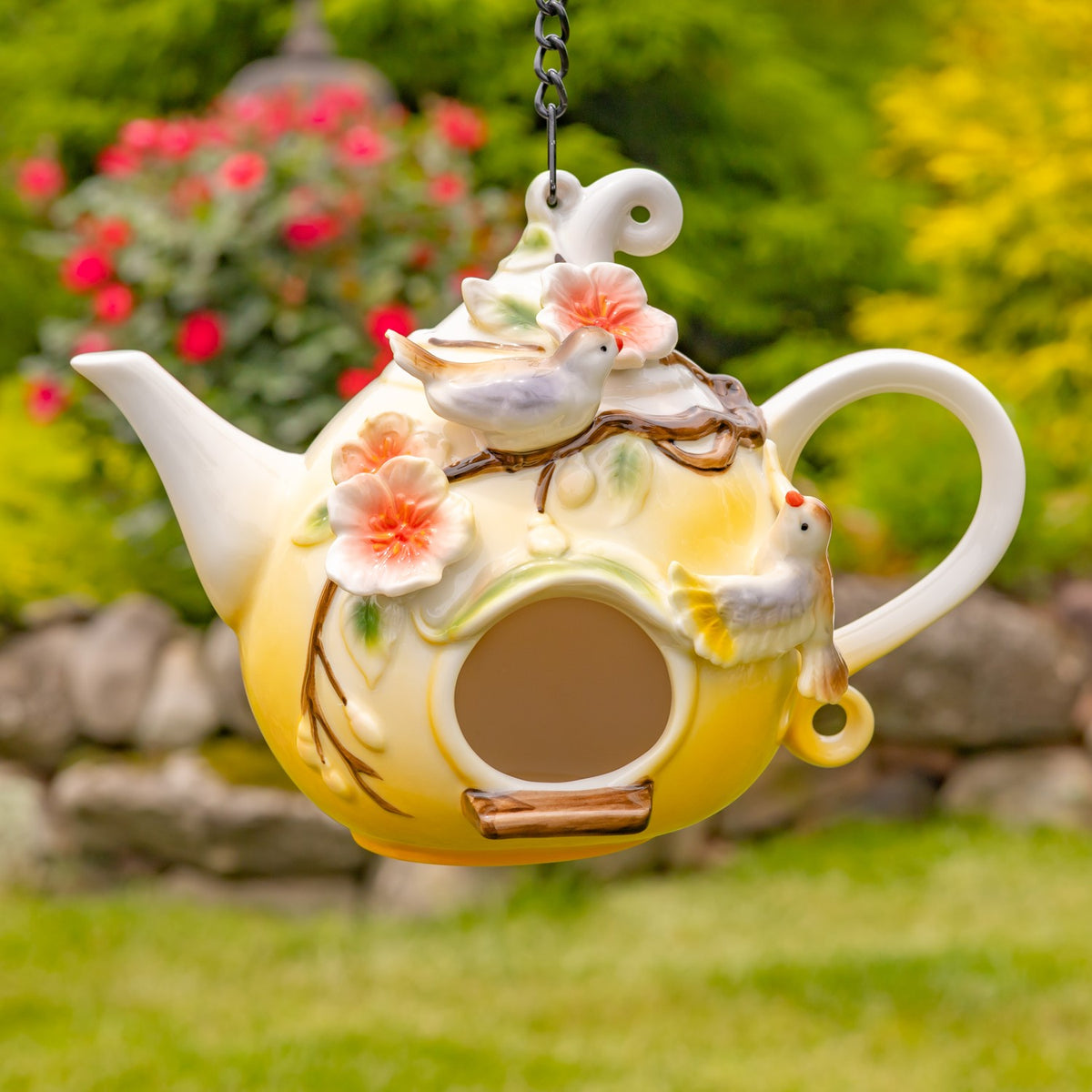 Yellow teapot birdhouse decorated with small birds and peach flowers, suspended outdoors