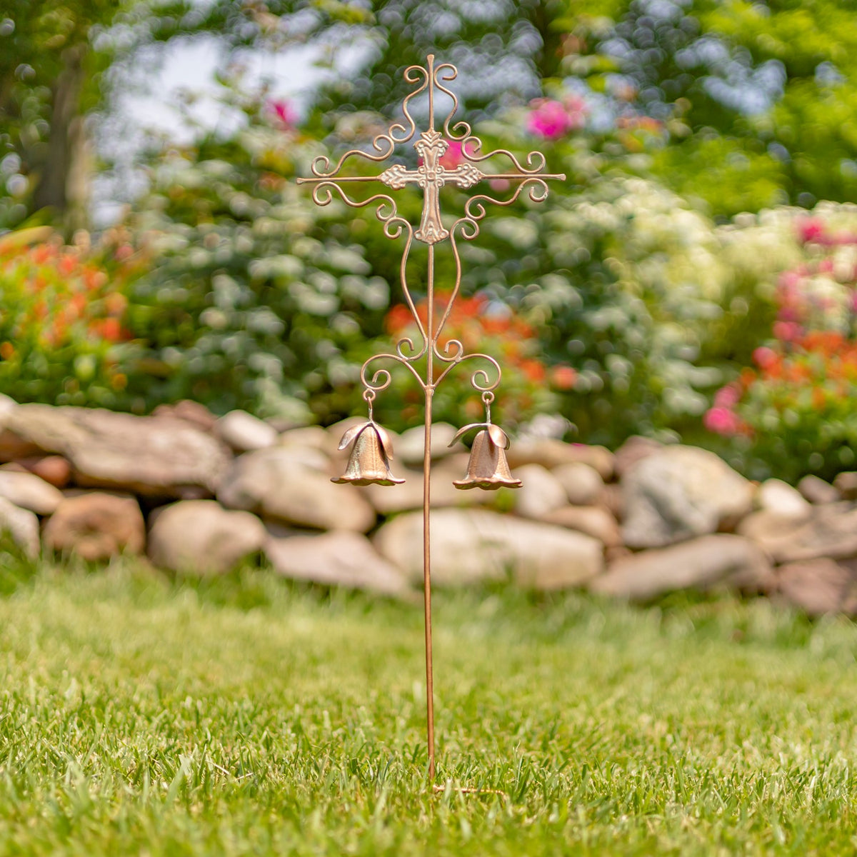 Slender ornate cross stake with symmetrical scrollwork and two bells displayed in a garden.