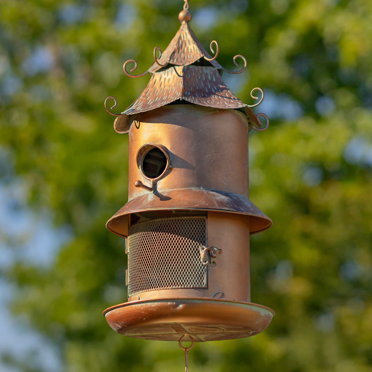 Hanging birdhouse feeder with a curled decorative roof, round entrance, mesh seed compartment, and bells below.