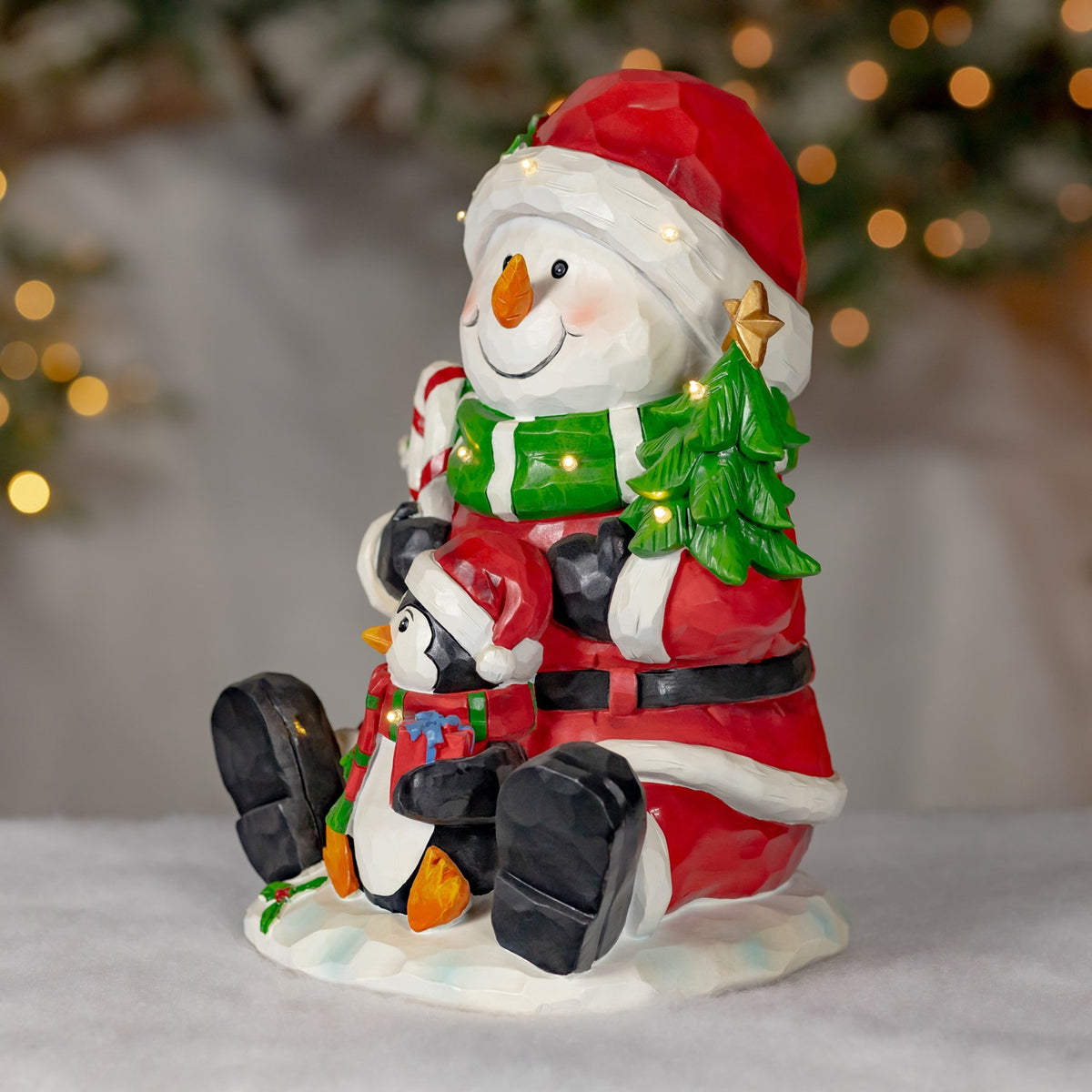 Angled view of the seated snowman and penguin, showing the snowman's red coat and the tree at his side.