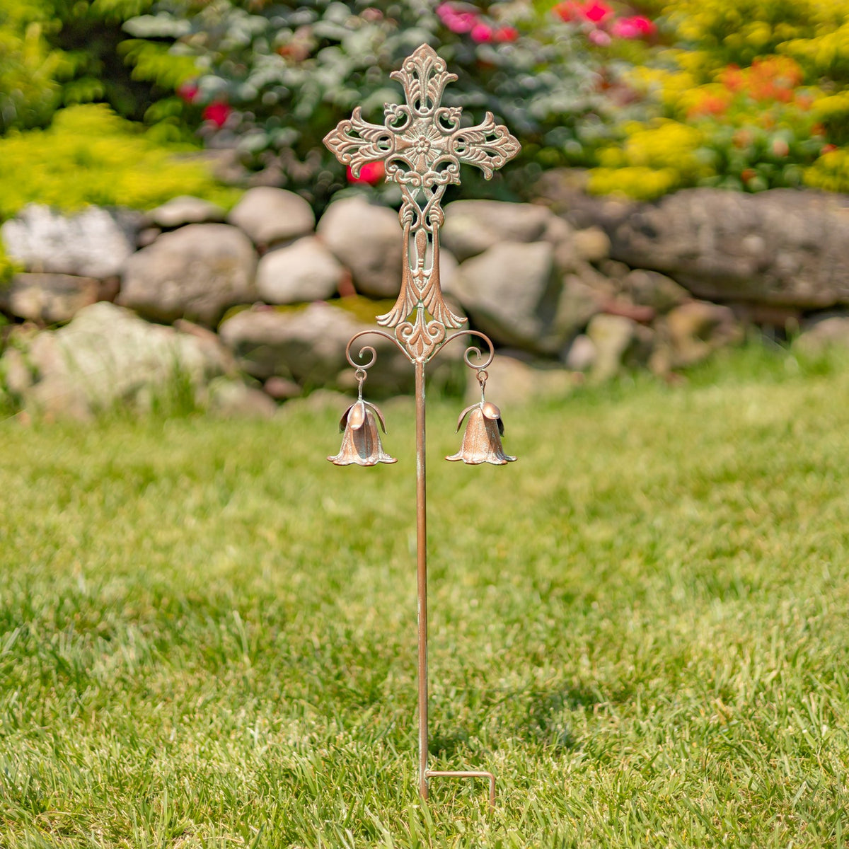A pale, scrolling cross stake with two dangling bells stands alone in the lawn.