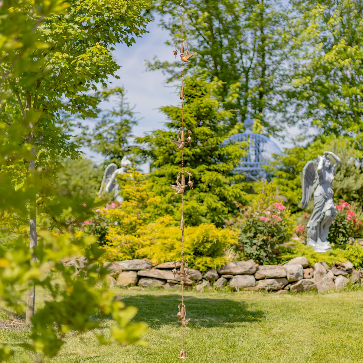 The long ornament chain hangs in a garden, with several birds and bells visible above a stone wall.