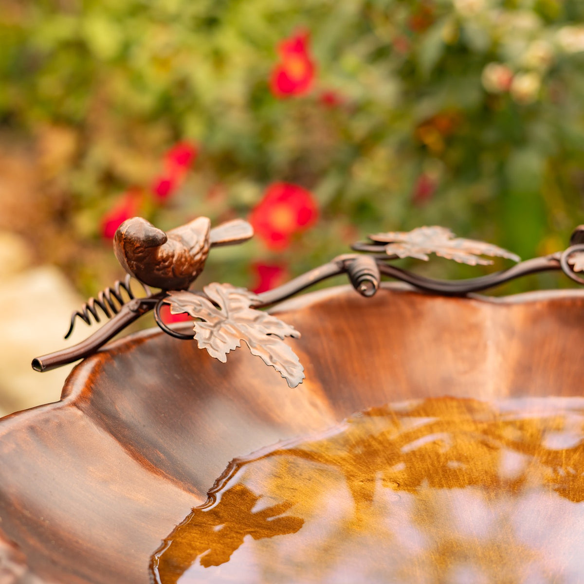 Close-up of a small bird and leaf ornament on the birdbath rim above the reflective water.